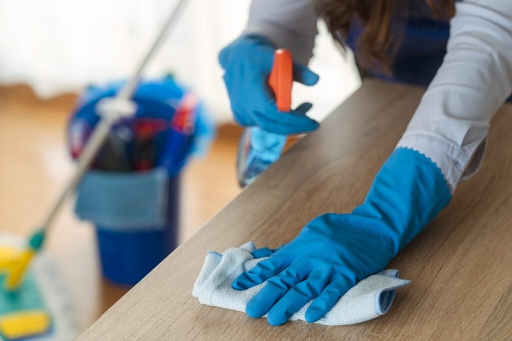 A person wearing blue gloves cleans a wooden surface with a white cloth, with a bucket of janitorial services in Baton Rouge supplies and a mop in the background.