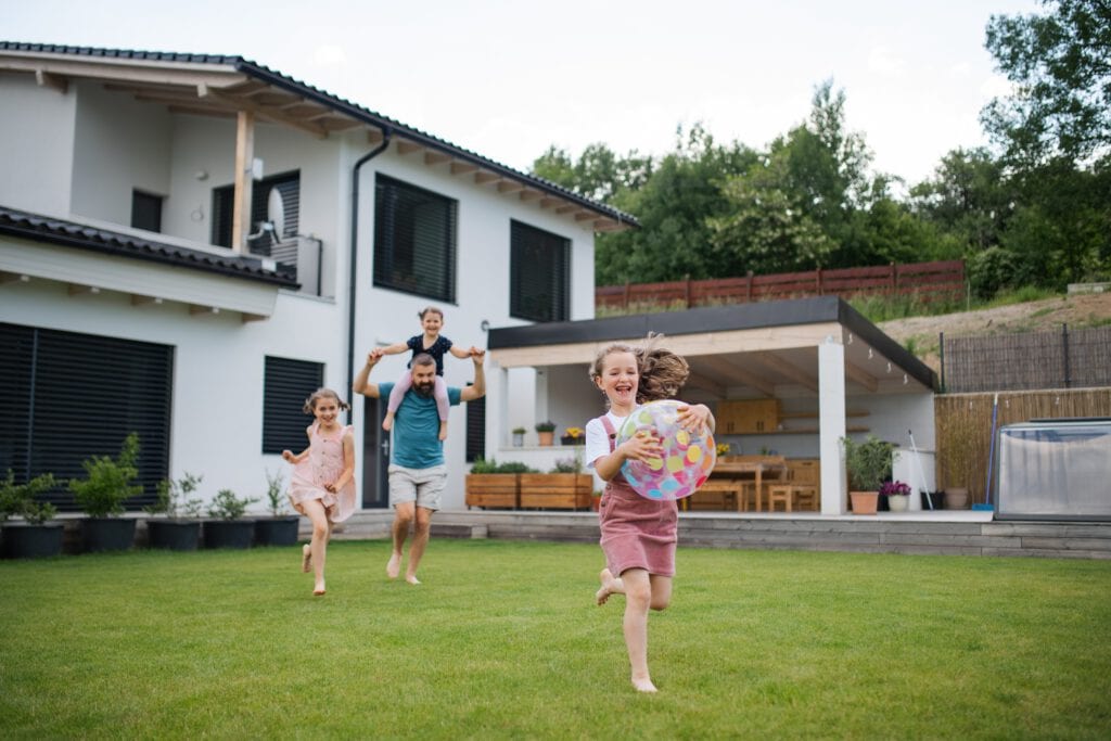 A family with two children plays in the backyard of a modern home. The kids are running on the grass, one with a toy, followed by their smiling parents who have recently utilized residential cleaning in Baton Rouge to maintain their pristine yard.