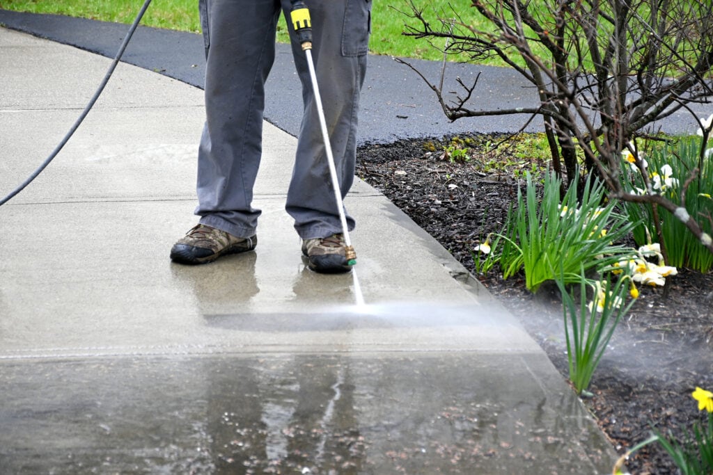 A person engaged in industrial cleaning, pressure washing a concrete sidewalk in Baton Rouge, focusing on where the nozzle sprays water. The scene includes green grass and blooming daffodils.