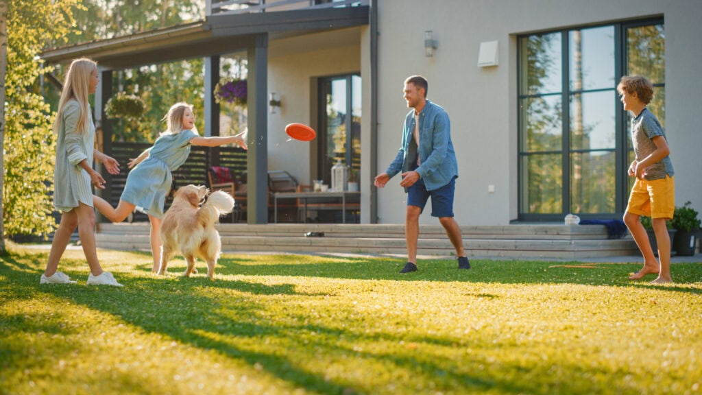 A family of four and their dog playing with a frisbee on a sunny backyard lawn. The house, benefiting from janitorial services in Baton Rouge, has large windows under a clear blue sky.