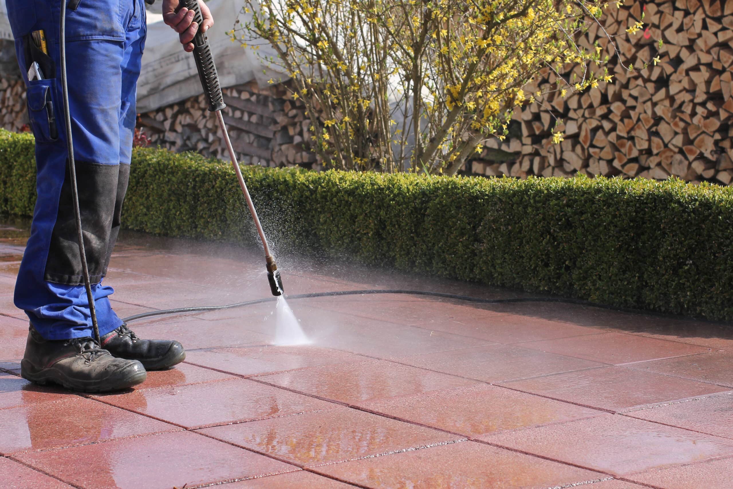 A person in blue jeans and work boots uses a high-pressure water spray to clean a red tiled patio, with neatly stacked wood and a green hedge in the background as part of a residential cleaning service in Baton Rouge.