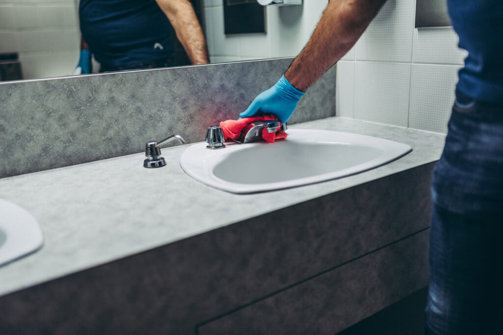 A person wearing blue gloves performs janitorial services in Baton Rouge, cleaning a white sink with a red cloth in a bathroom, with a mirror reflecting their torso.