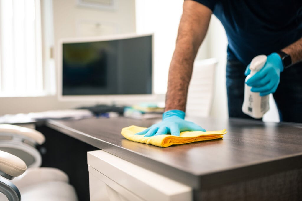 A person wearing blue gloves performs an office cleaning in Baton Rouge on a dark wood countertop with a yellow cloth, holding a spray bottle. A blurred background shows a modern room with a television.