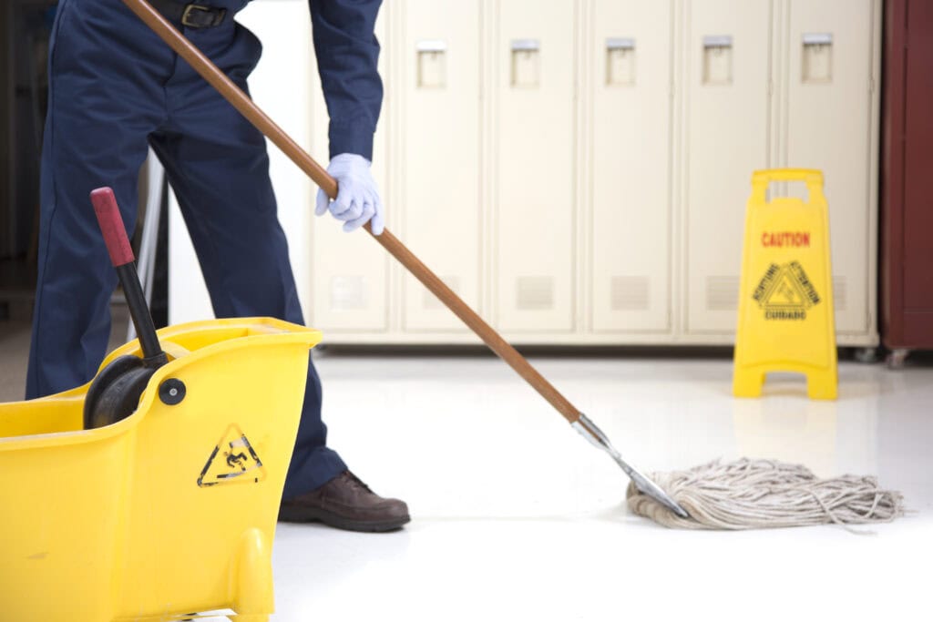 A janitor in a blue uniform and gloves mops a school corridor with a yellow mop bucket and caution sign nearby, indicating a wet floor. This scene exemplifies the high-quality janitorial services in Baton Rouge.