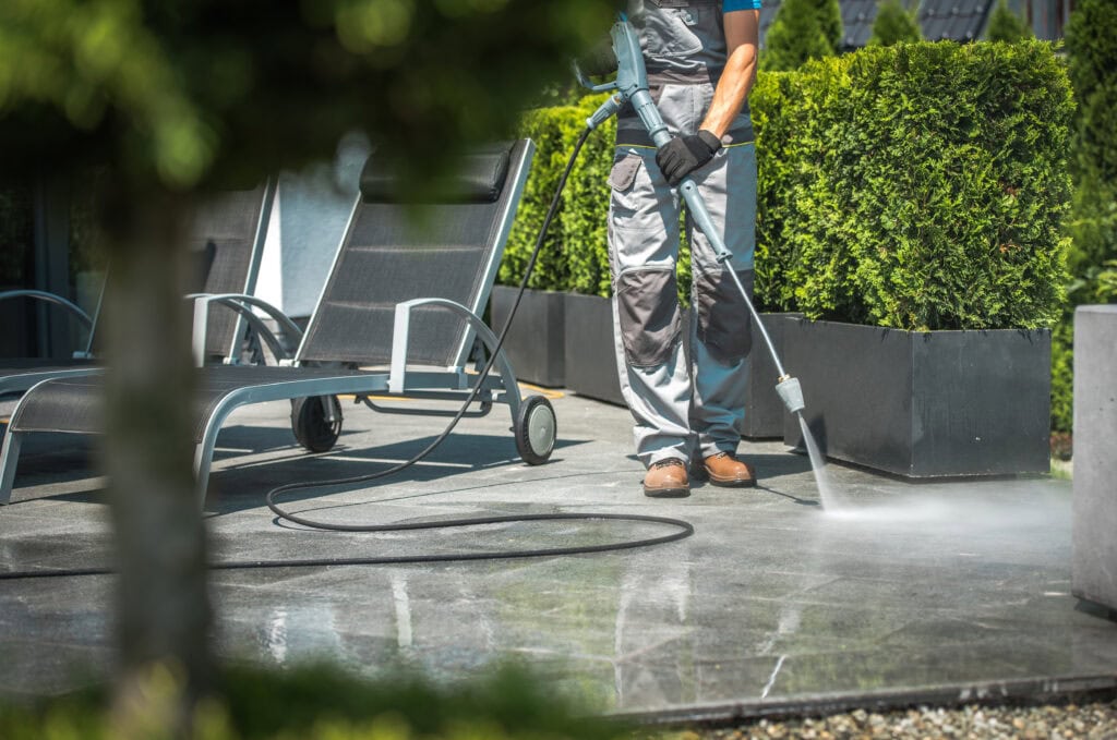 A person cleaning a patio using a high-pressure washer, surrounded by garden furniture and lush greenery, on a sunny day in Baton Rouge.