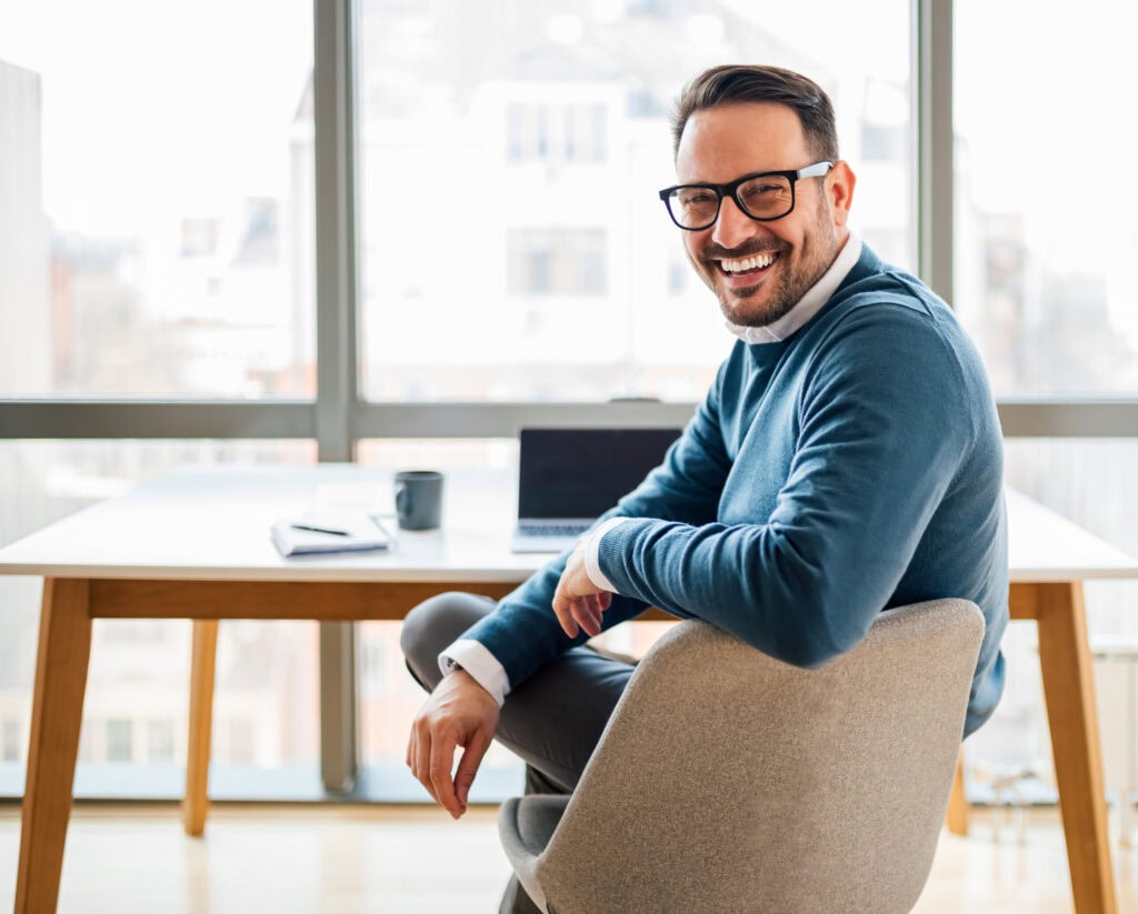 A cheerful man wearing glasses and a blue sweater sits on a chair at a modern office desk, serviced by janitorial services in Baton Rouge, looking at the camera with a bright smile, with a laptop and phone nearby. Large windows in the background.