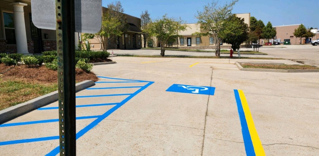 Two accessible parking spaces with blue and white wheelchair symbols, marked by yellow lines, in a sunny parking lot near a commercial building in Baton Rouge.