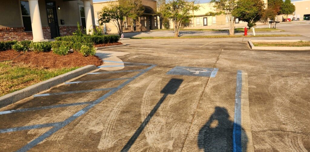 A parking lot with designated blue parking lines and a wheelchair symbol, with shadows stretching across from low-hanging sunlight. A building with columns, maintained through industrial cleaning in Baton Rouge, is visible on the left.