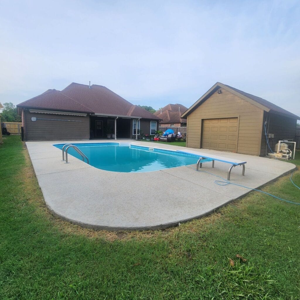 A front and backyard view showcases a house with a swimming pool. There's a concrete deck around the pool with two diving boards, and a grassy area bordering the concrete. A detached garage or shed with a single door is to the right of the pool. The sky is partly cloudy.