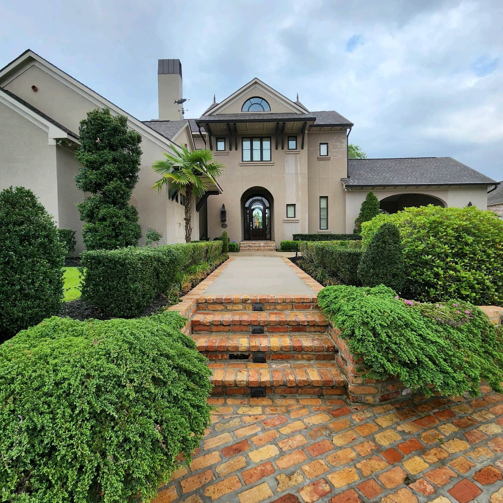 A two-story, modern gray house with a gabled roof and tall chimney stands proudly. The front yard features manicured hedges, lush green bushes, and a brick pathway leading to the entrance. The sky is partially cloudy.