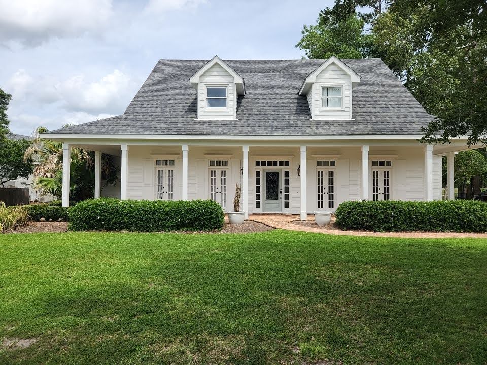 A charming white home with a steep gray roof and two dormer windows adorned with shutters. The front porch features four white columns and multiple French doors. A manicured lawn and lush green hedges, maintained through soft washing, enhance the welcoming appearance of the property.