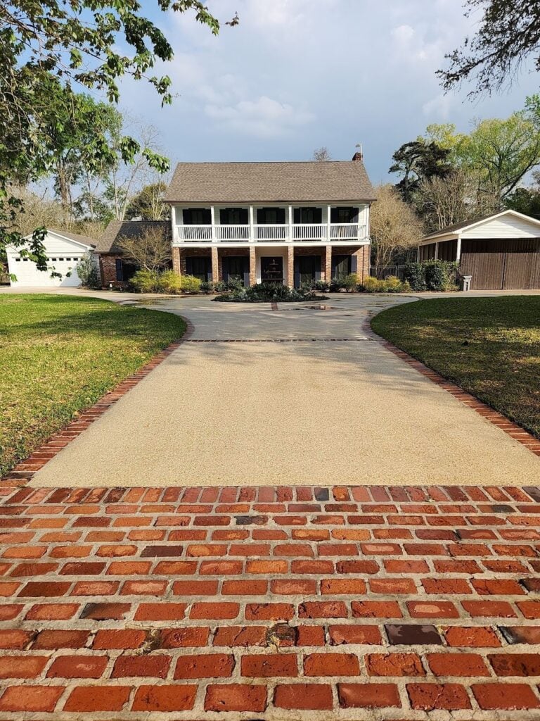 A two-story house with a concrete driveway leading to it. The house features a front porch with white railings and pillars, a balcony above the porch, and is surrounded by trees and shrubs. Recently pressure washed, the home sparkles under the partly cloudy sky.