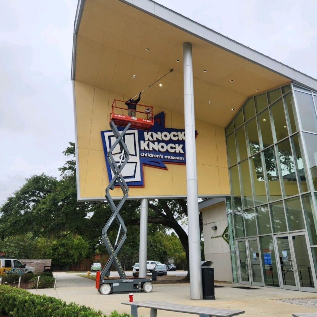 A worker on a scissor lift adjusts fixtures near the sign of the Knock Knock Children's Museum, which features glass windows and a modern architectural design—an example of prime commercial space. Trees and maintenance vehicles are in the background.