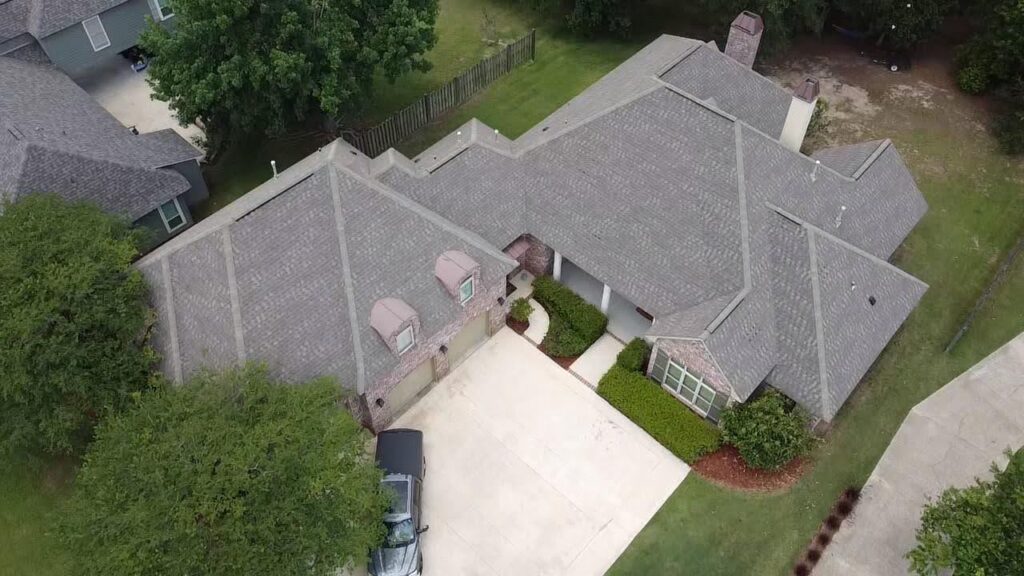 Aerial view of a suburban home with a well-maintained grey roof, surrounded by trees and greenery. The house features a large driveway with a car parked and a neat lawn, suggesting regular roof maintenance. Adjacent houses and a fenced backyard are also visible.