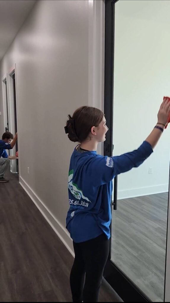 Two individuals are performing move-out cleaning on glass doors and windows in an indoor hallway. The person in the foreground, wearing a blue shirt and a black bow in their hair, is wiping a full-length glass door. Another person is seen in the background cleaning a window.