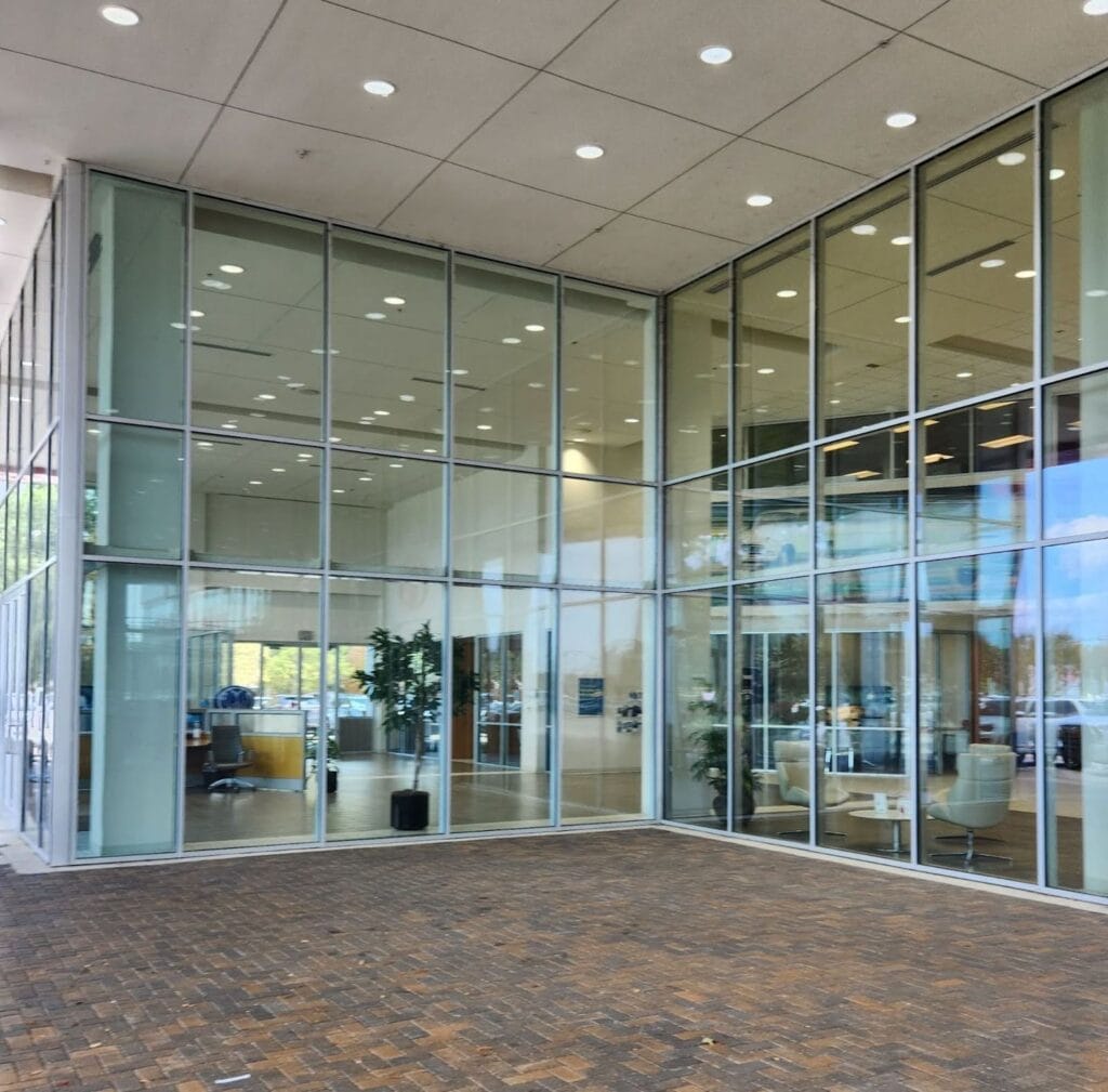 A modern office building with large glass windows reflecting the outdoor scene, showcasing an interior with white walls, potted plants, and seating areas. The pristine windows highlight the cleanliness of the space. The exterior features a brick-paved walkway under a ceiling with recessed lights.