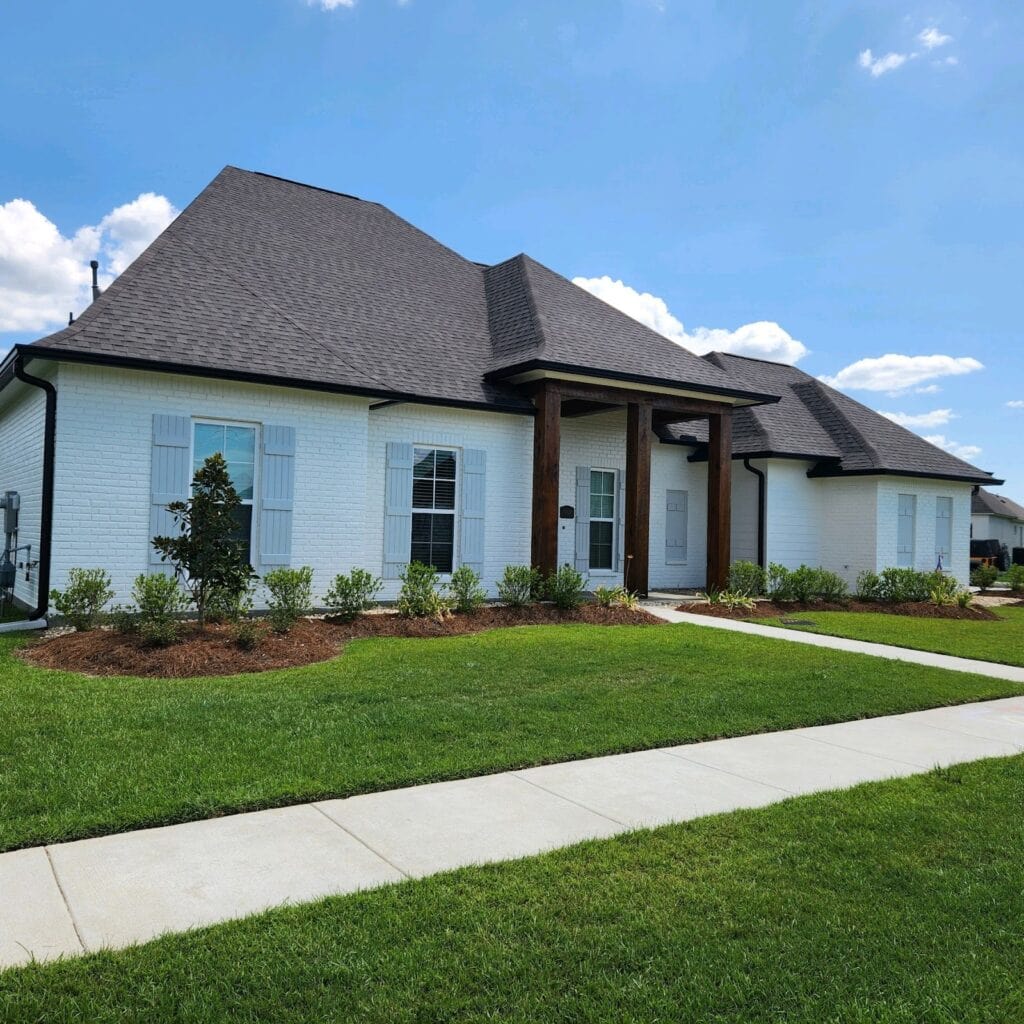 A charming piece of residential real estate, this suburban house boasts a white brick exterior, black roof, and wooden pillars on the porch. The property features well-maintained lawns, trimmed hedges, and a paved walkway under a clear sky with a few fluffy clouds.