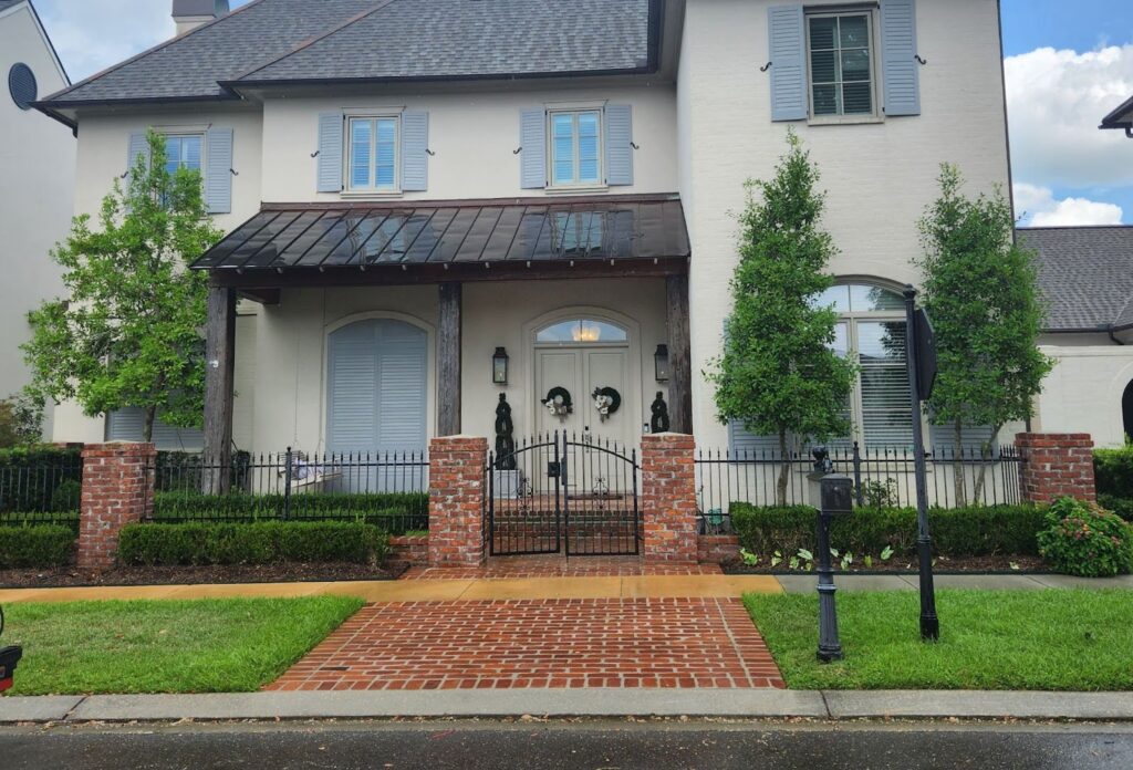 A two-story house with a gray roof and light-colored exterior, featuring blue shutters and a large front porch supported by brick columns. The yard, meticulously maintained with soft washing techniques, showcases trimmed bushes, two trees, and a brick pathway leading to a black gate. The sky is partly cloudy.