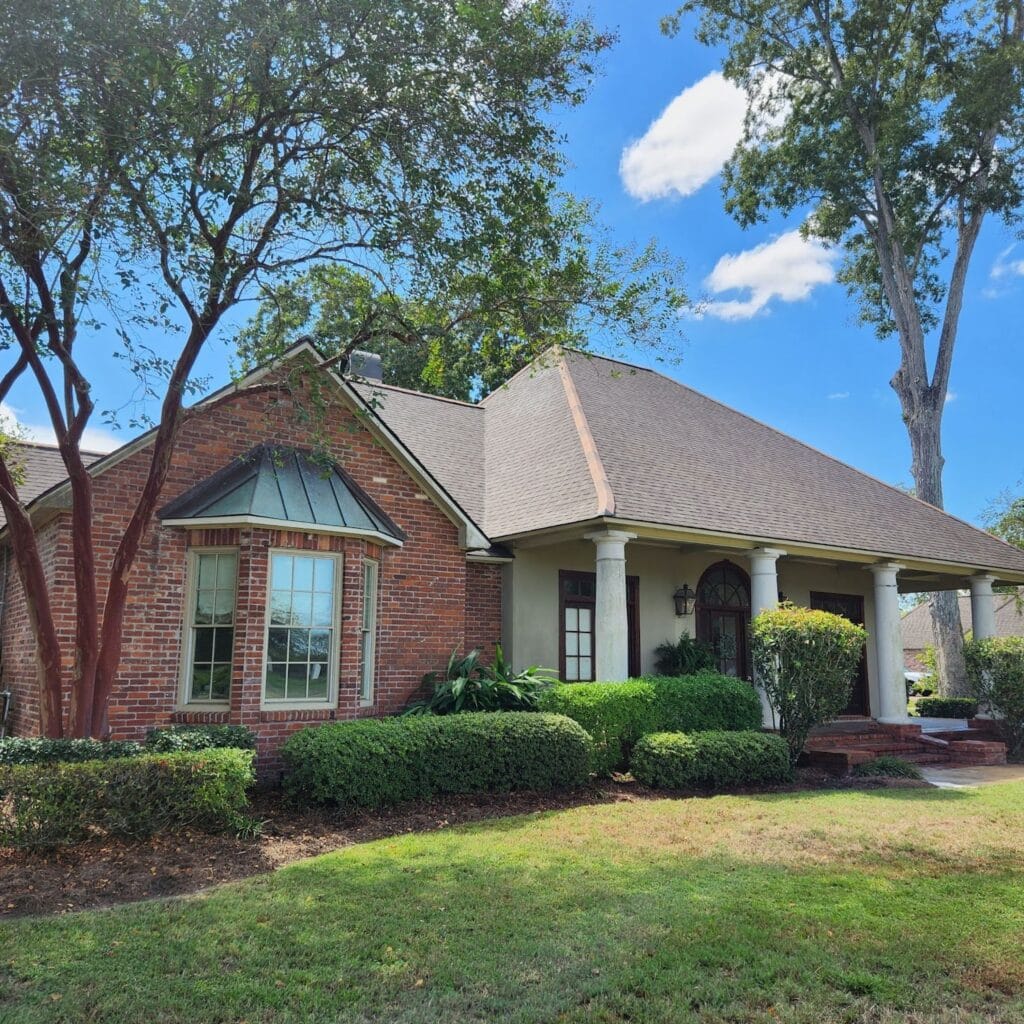 A brick house with a steeply pitched roof features a large bay window, white pillars framing the front porch, manicured shrubs, and a green lawn. The soft washing of its exterior highlights its charm. Tall trees with green foliage stand in the background against a clear blue sky dotted with a few white clouds.