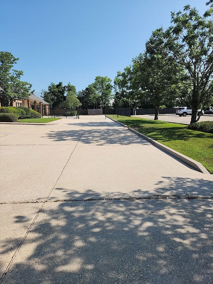 A wide, sunlit driveway extends forward, flanked by trimmed green lawns and trees casting shadows. Houses are visible on the left, and a white truck is parked on the right. A person is seen walking down the front driveway in the distance. The sky is clear and blue.