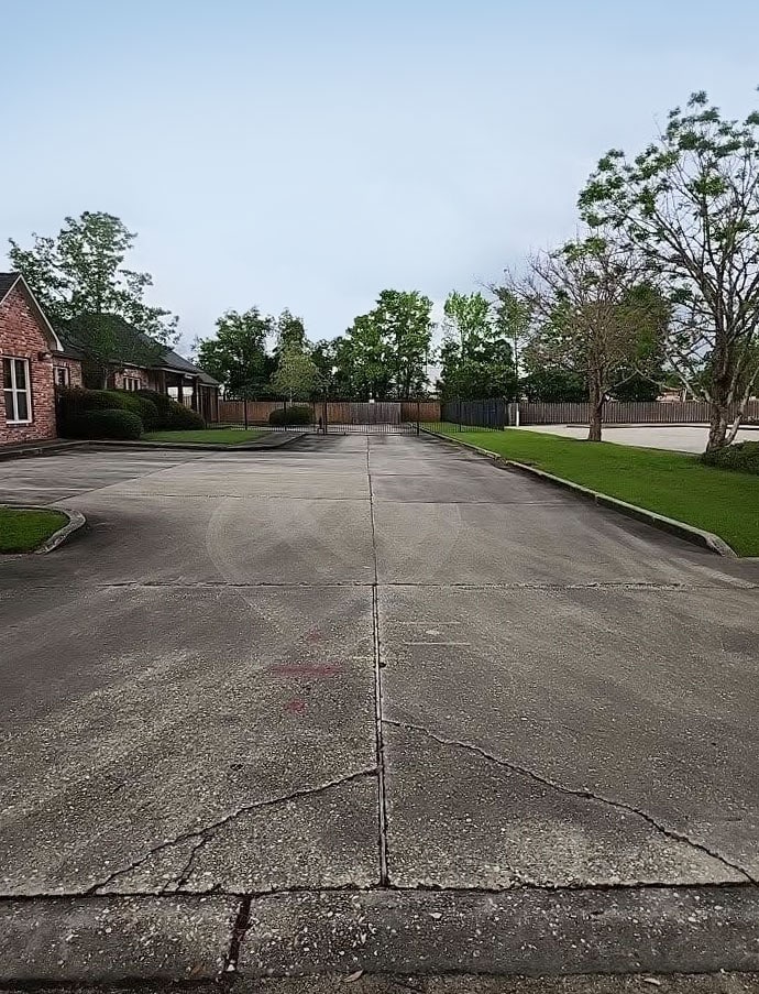 An empty residential street with a concrete pavement featuring visible cracks, flanked by brick houses, green lawns, and several trees. The sky above is clear. This quaint setting could serve as the perfect first impression for a website front depicting suburban tranquility, ending at a wooden fence in the background.