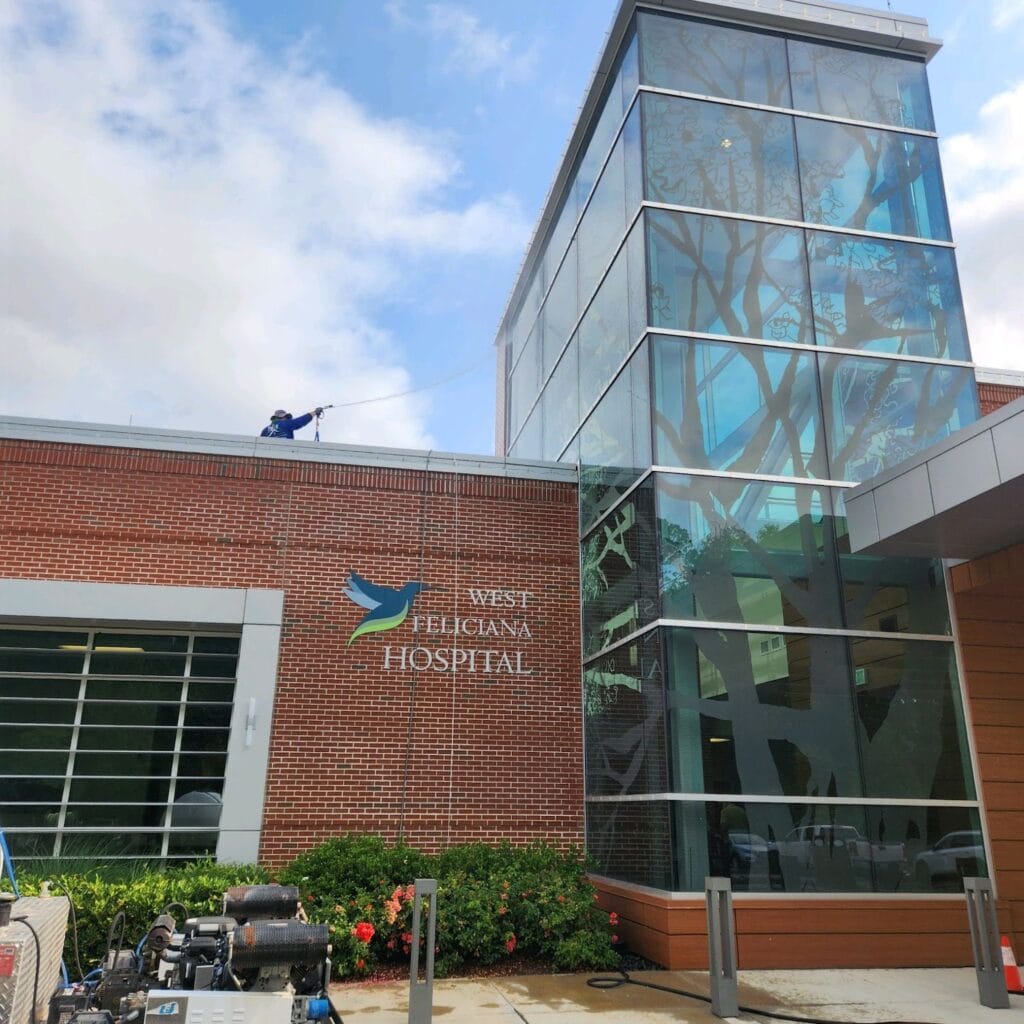 A worker on the roof of West Feliciana Hospital uses a power washer for professional cleaning of the building's exterior. The hospital has a modern glass and brick facade, with its name and logo visible on the front. Lush greenery and equipment are present at the entrance.